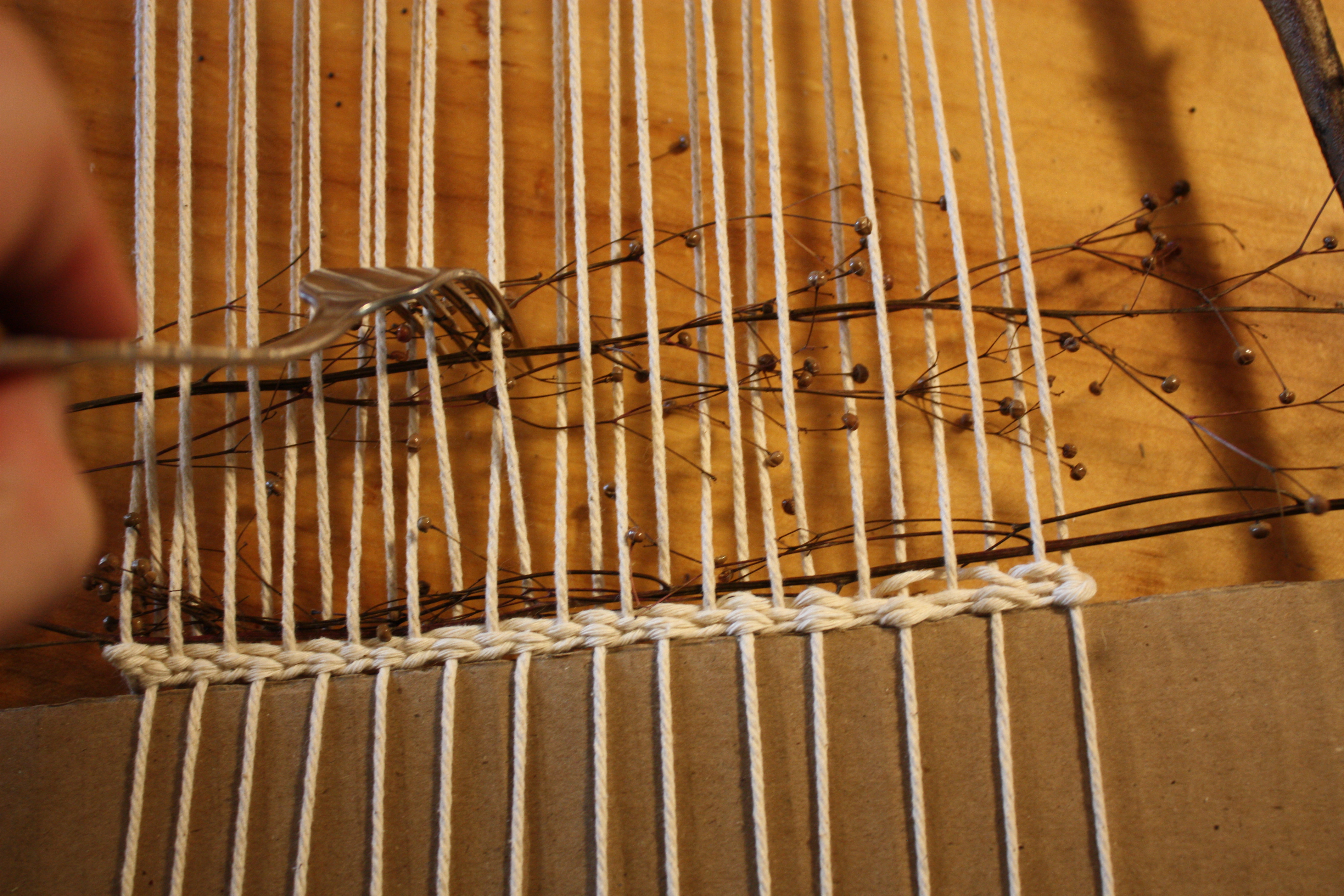  In-progress weaving with natural materials — grasses, twigs, flowers layered between rows of yarn.