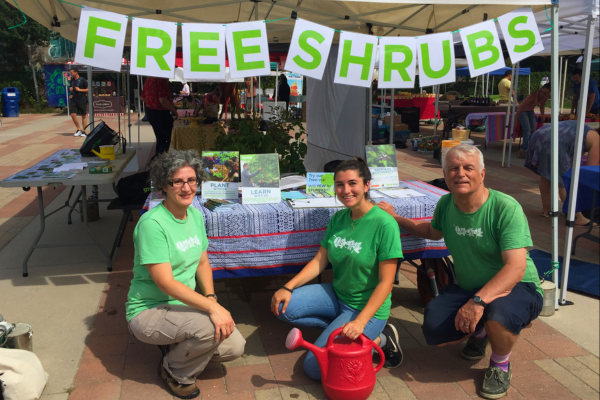 Three LEAF volunteers in front of an outreach table, at an outdoor farmer’s market. A sign above them reads “Free shrubs”