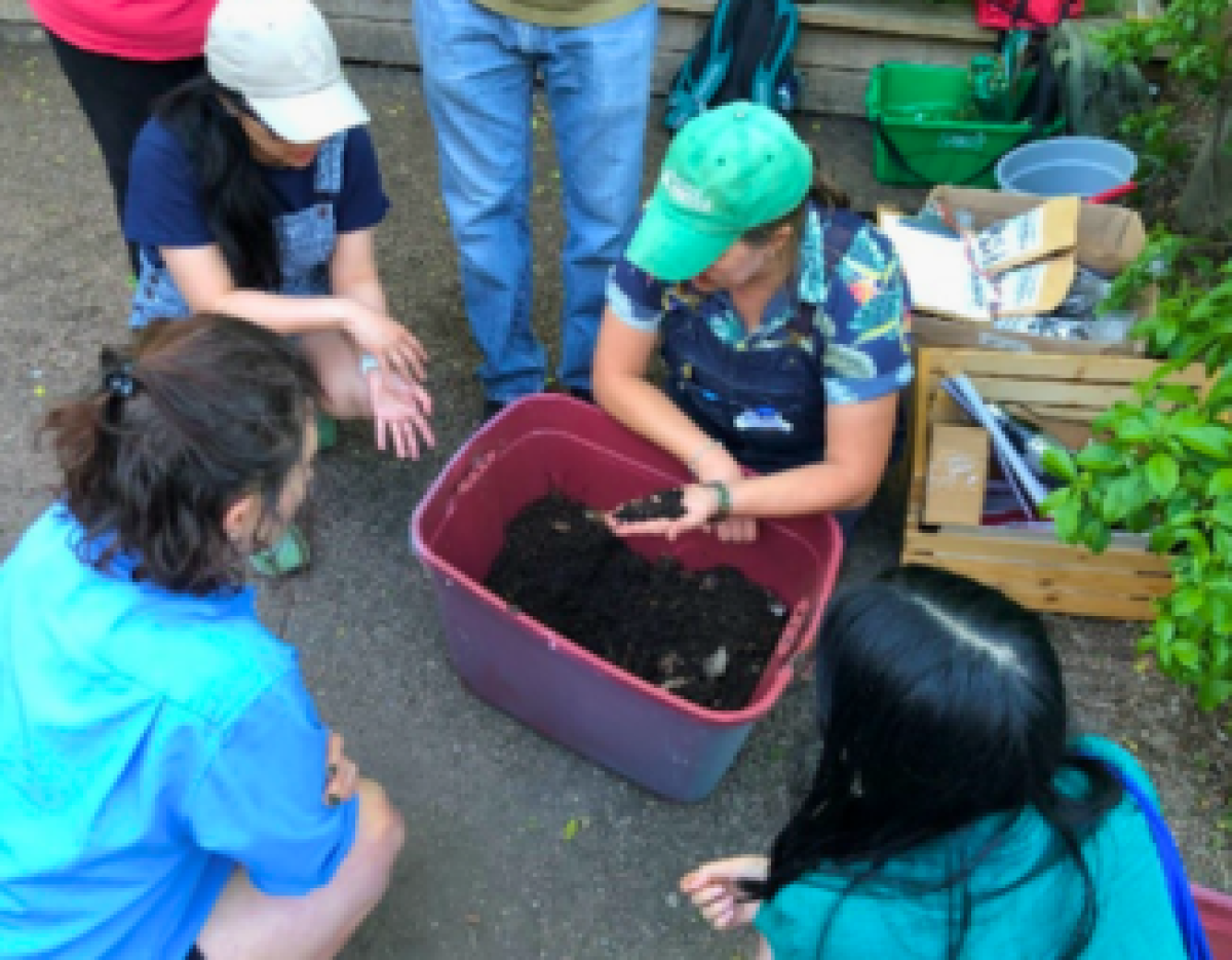 ©2026 Sarah Halonen/LEAF A group of volunteers and workshop leader crouched around a red bin filled with garden soil.