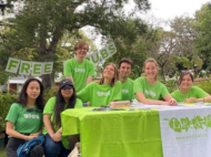 A group of 7 people in green LEAF shirts are sitting at a table smiling at the camera after giving t