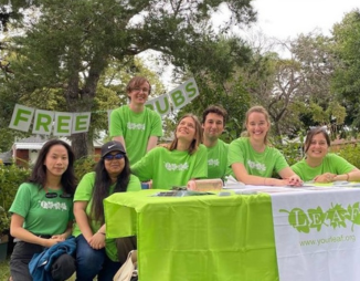 A group of 7 people in green LEAF shirts are sitting at a table smiling at the camera after giving t